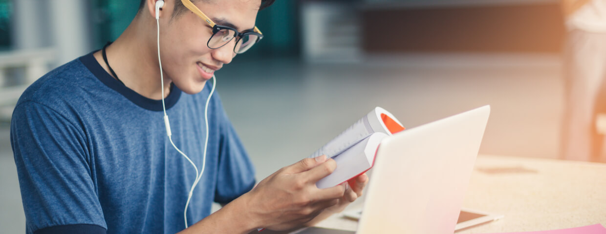 student boy reading book for exam and listening to music via headphones with laptop.