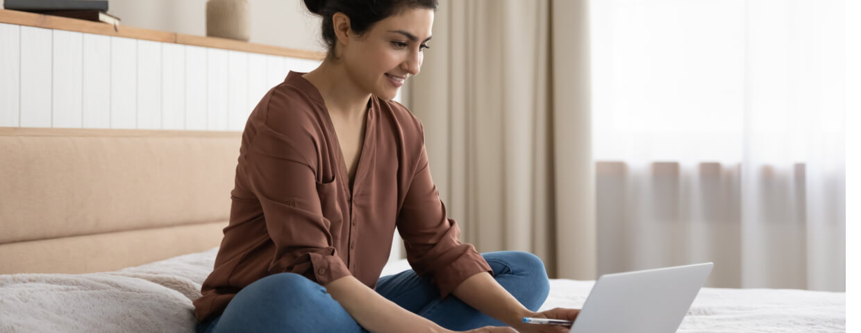 online student sitting on a bed with laptop and pen