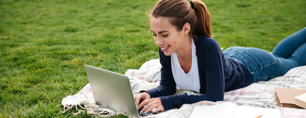 smiling teen student girl doing homework on laptop computer while lying on the grass at park, summer school