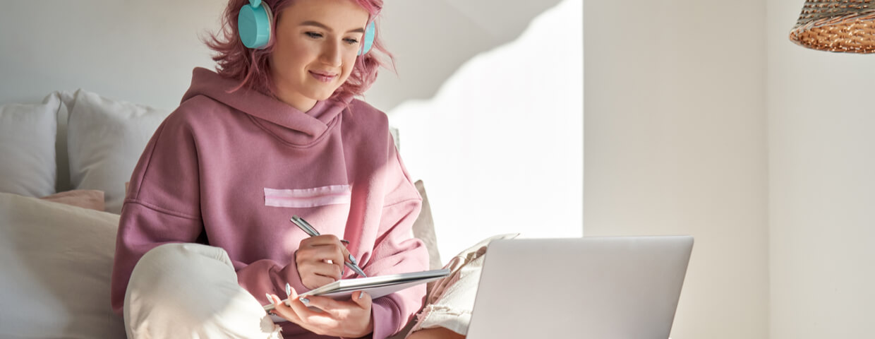 teenage girl with pink hair wearing headphones doing homework or online school on a laptop while writing notes