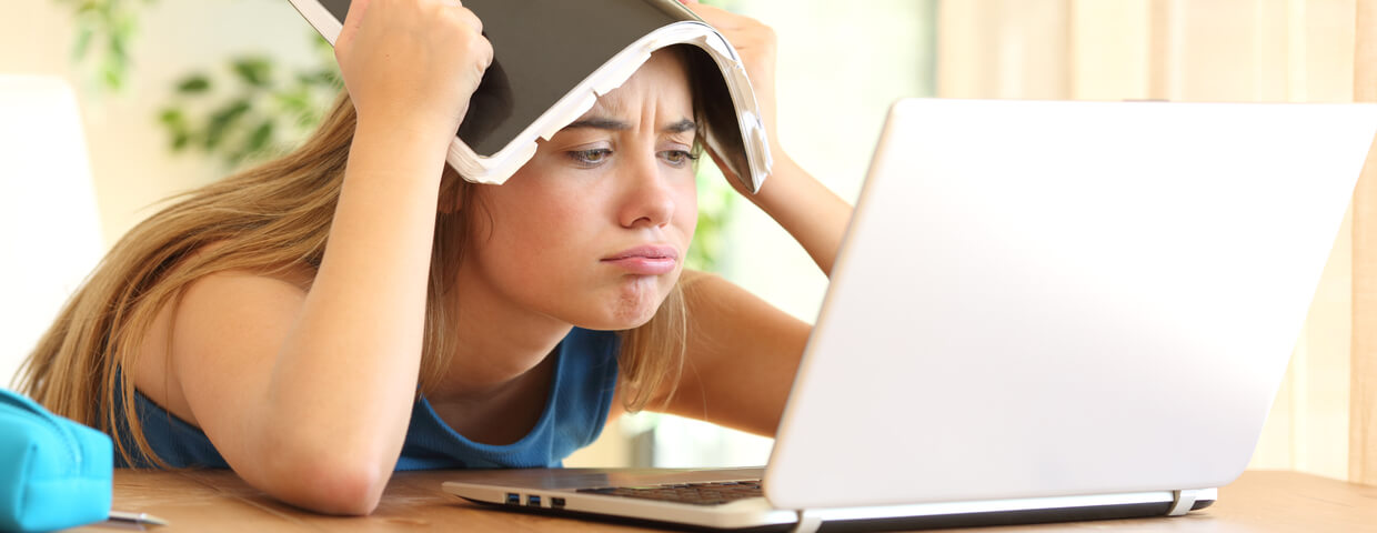 teen student struggling with online school, notebook on her head at a table at home in front of a laptop
